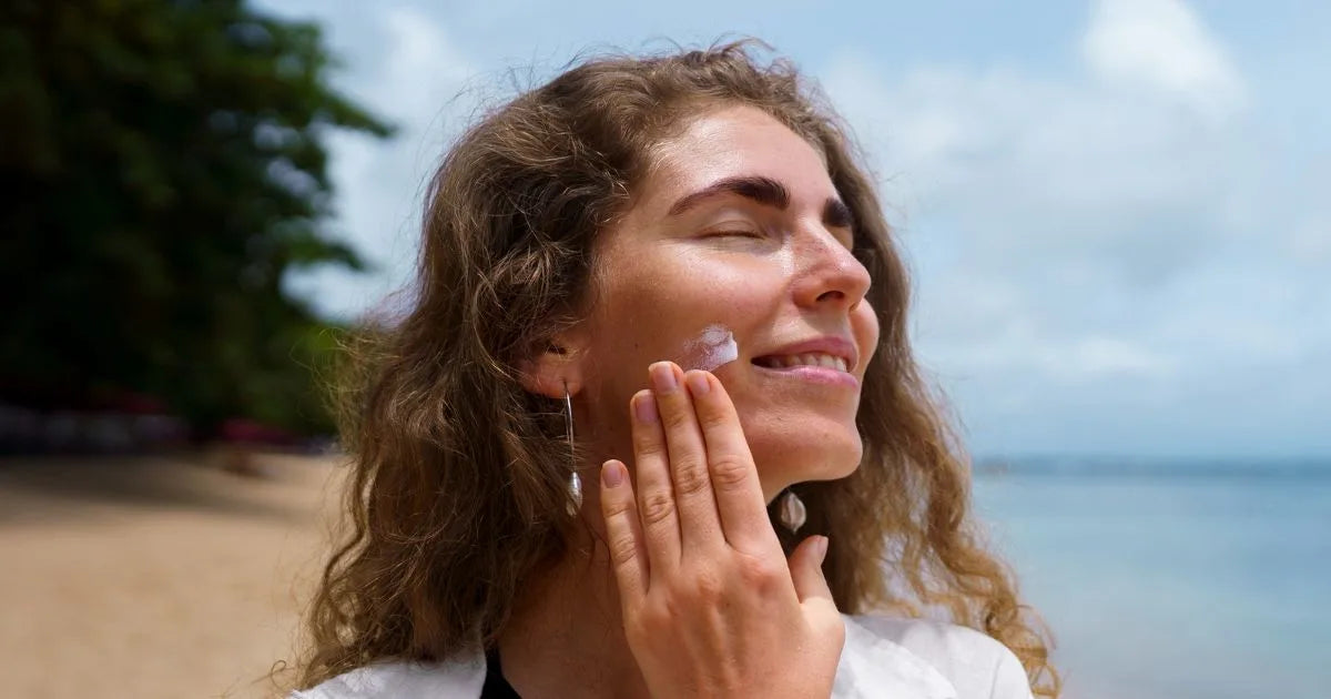 Smiling woman on a beach applying cream to her cheek in the sunshine